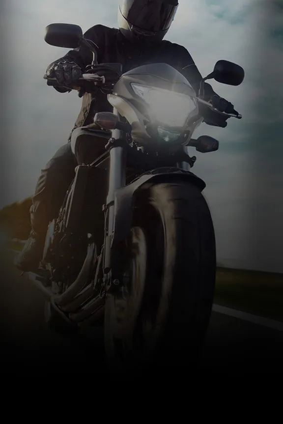 Frontansicht eines Motorradfahrers mit Helm und leuchtendem Scheinwerfer auf einer Landstraße bei bewölktem Himmel.